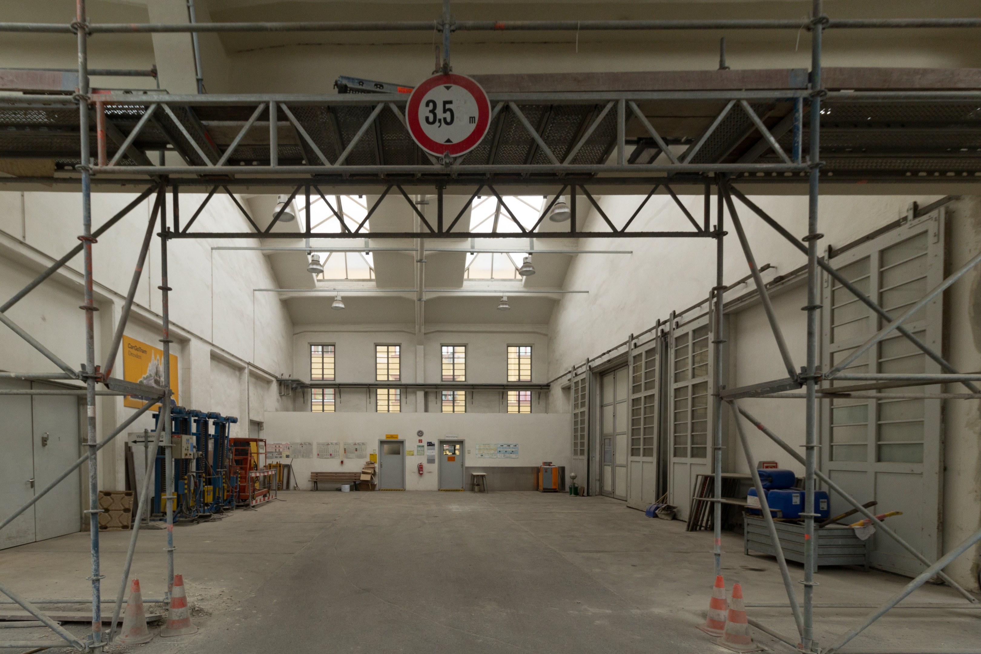 Lagerhalle mit ausgedehnten Gerüsten, Verkehrszeichen, Metallstangen, einem Schild, Maschinen, einer Tür, Fenstern, Wänden, Deckenleuchten und einem Dach.