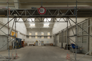 Lagerhalle mit ausgedehnten Gerüsten, Verkehrszeichen, Metallstangen, einem Schild, Maschinen, einer Tür, Fenstern, Wänden, Deckenleuchten und einem Dach.