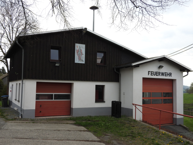 Feuerwache mit einem roten und weißen Gebäude, Straße, Gras, gelben Blumen und umliegenden Elementen unter einem Himmelhintergrund.