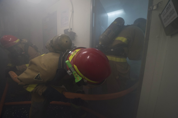 Feuerwehrleute in Schutzausrüstung mit Sauerstoffflaschen betreten einen raucherfüllten Raum durch eine Tür, mit Rohren und einer Lampe im Hintergrund.