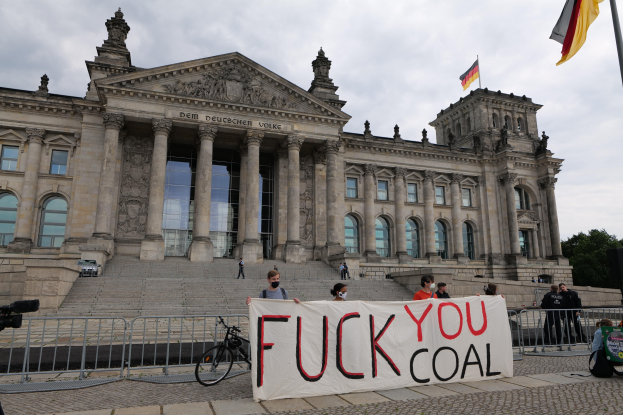 Protestierende halten ein "Fuck You Coal"-Schild vor dem Reichstaggebäude in Berlin, Deutschland, mit seinen architektonischen Details, einem Fahrrad, Bäumen, einem Fahnenmast und einem bewölkten Himmel.