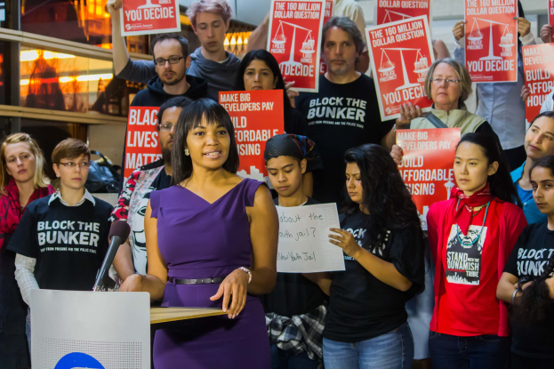 Eine Black Lives Matter-Aktivistin spricht vor einem Publikum mit Mikrofon an einem Rednerpult, während das Publikum Schilder hält, mit Lichtern und Gegenständen im Hintergrund.