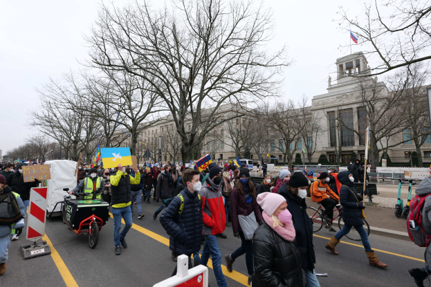 Eine große Gruppe von Menschen marschiert auf einer Straße in Washington, D.C. und hält Schilder und Banner hoch, während einige Fahrräder fahren, unter einem klaren blauen Himmel.