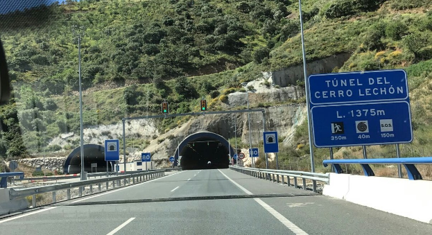 Ein Auto, das durch einen Tunnel neben einer Straße mit Geländern, Schildern, Pfählen, Verkehrszeichen, Bäumen, Felsen und einem klaren blauen Himmel fährt.