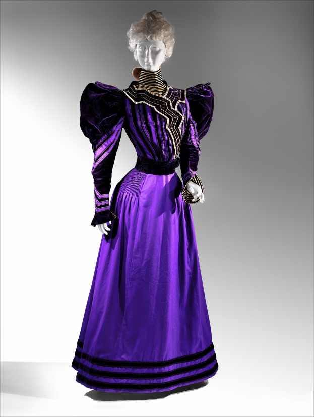 Eine Schaufensterpuppe in einem lila Abendkleid mit schwarzem und goldenem Muster, das eine eng anliegende Taille, einen langen Rock mit goldener Stickerei und Perlen und eine Spitzenverzierung an der Taille hat, kombiniert mit einem eleganten Dutt und zarten Ohrringen.