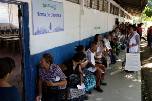 Eine Gruppe von Menschen sitzt auf Bänken vor einem Gebäude, wartend in der Schlange, um an einer Wahllokal in Guadalajara, Mexiko, mit einer Texttafel an der Wand und einer Tür links und Bäumen im Hintergrund zu wählen.