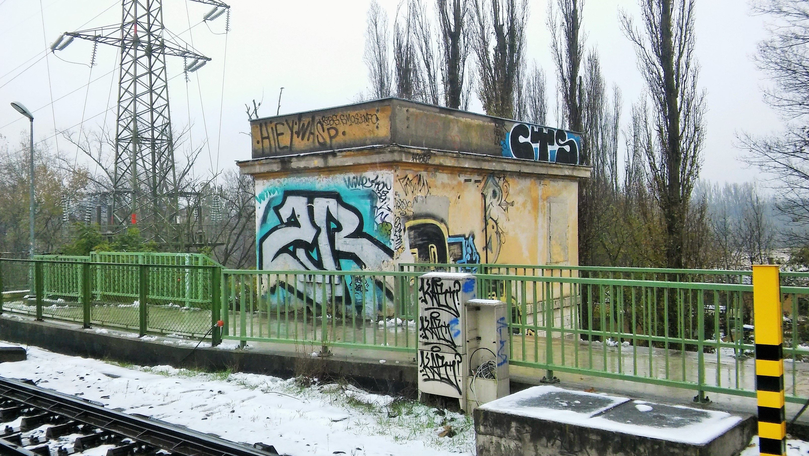 Eisenbahnschiene mit Graffiti an der Seite, umgeben von Geländern, Pfählen, Lampen und Bäumen, mit Türmen und Drähten im Hintergrund unter einem klaren Himmel und Schnee am Boden.