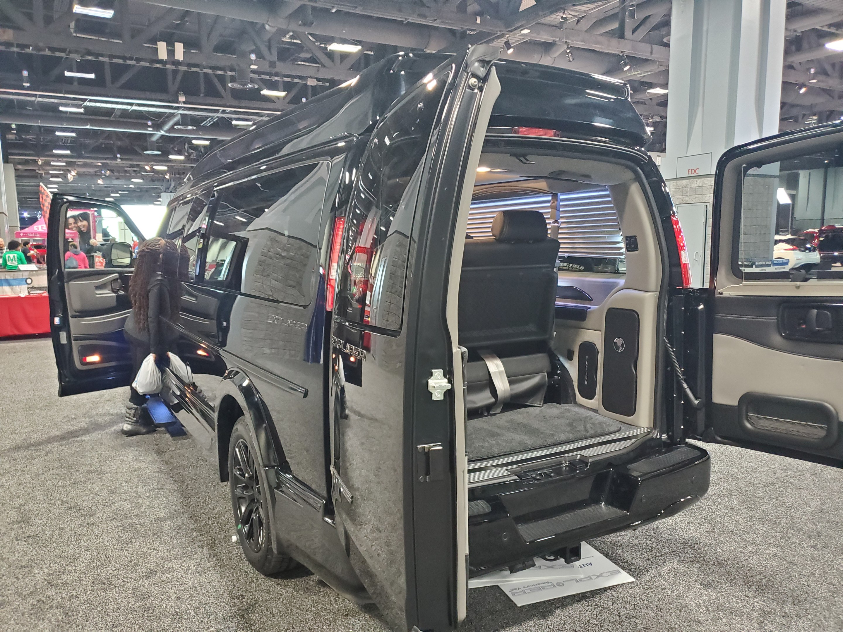 Schwarzer Mercedes-Benz Sprinter Van mit geöffneten Türen auf einer Ausstellung; Heck mit einem Tisch mit Gegenständen, einer Fahne mit Text und einem Dach mit Deckenlampen und Metallstäben.
