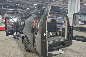 Schwarzer Mercedes-Benz Sprinter Van mit geöffneten Türen auf einer Ausstellung; Heck mit einem Tisch mit Gegenständen, einer Fahne mit Text und einem Dach mit Deckenlampen und Metallstäben.