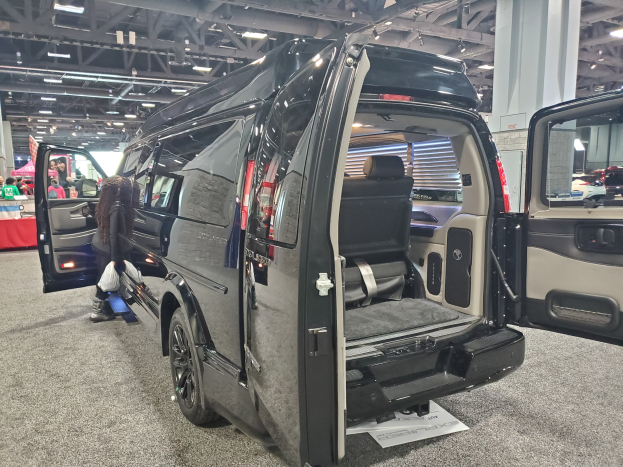 Schwarzer Mercedes-Benz Sprinter Van mit geöffneten Türen auf einer Ausstellung; Heck mit einem Tisch mit Gegenständen, einer Fahne mit Text und einem Dach mit Deckenlampen und Metallstäben.