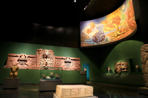 Ein Museumsraum mit verschiedenen Artefakten, darunter Skulpturen, Gegenstände auf Tischen, eine Texttafel, eine Wandmalerei und Deckenleuchten im Mexiko-Stadt-Museum der Anthropologie.