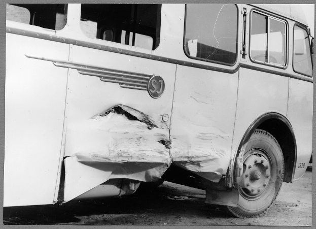 Ein Schwarz-Weiß-Foto eines beschädigten Busses am Straßenrand, dessen Front eingedellt ist, Fenster zerbrochen und die Fahrertür offen steht.