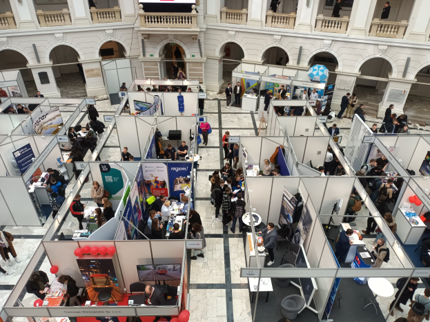 Große Gruppe von Menschen auf einer internationalen Messe mit Ständen, Bannern, Tischen, Stühlen und Ballons in einem Raum mit architektonischen Bögen, Säulen und einem Bildschirm im Hintergrund.