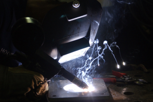 Ein Schweißer in einer dunklen Werkstatt verwendet einen Brenner, um an einem Metallstück zu arbeiten, wobei Werkzeuge auf einem nahen Tisch sichtbar sind.