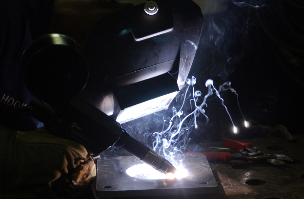 Ein Schweißer in einer dunklen Werkstatt verwendet einen Brenner, um an einem Metallstück zu arbeiten, wobei Werkzeuge auf einem nahen Tisch sichtbar sind.