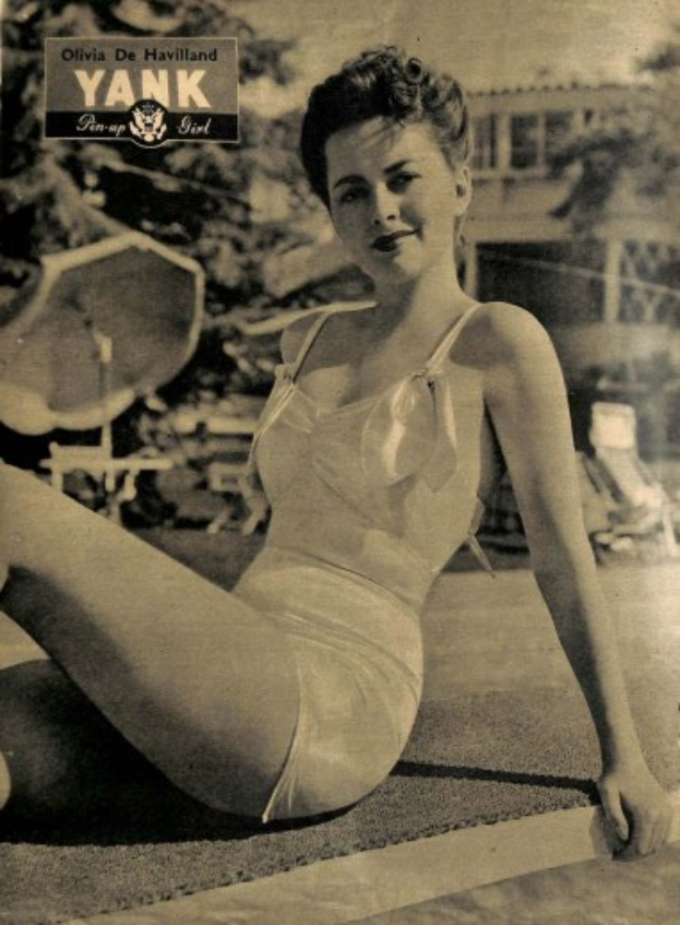 Olivia de Havilland in einem Badeanzug auf dem Boden sitzend und strahlend, mit Bäumen, Stühlen, einem Gebäude und Text im Hintergrund.