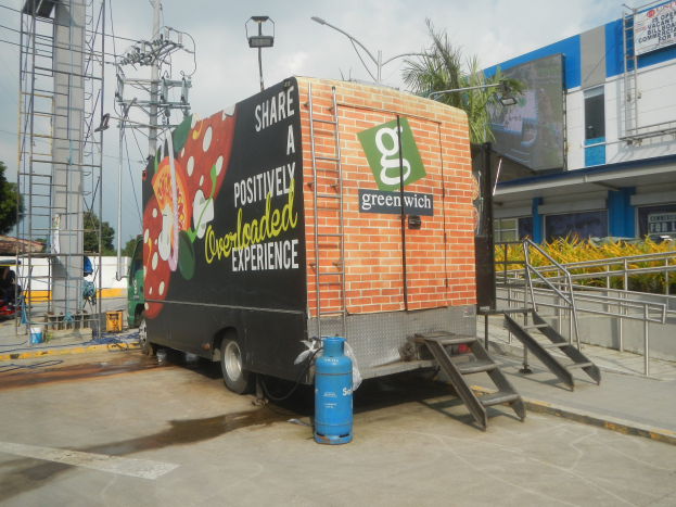Ein Lkw mit einer "Teile eine positiv überladene Erfahrung"-Aufschrift vor einem Gebäude geparkt, mit Stufen, einem Zylinder, Geländern, Strommasten, Laternen, Bäumen und einem bewölkten Himmel im Hintergrund.