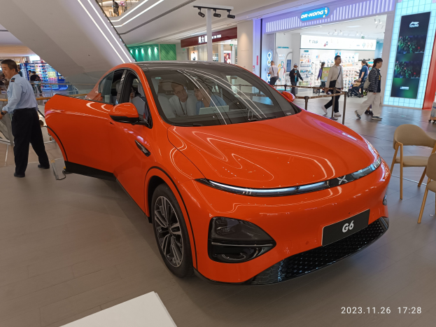 Orangefarbener Elektroauto in einer Ausstellungshalle umgeben von Menschen, mit Stühlen und einem Wagen rechts und Geschäften im Hintergrund.