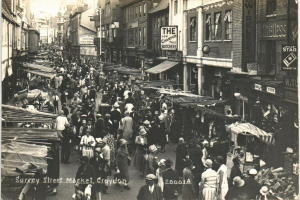 Ein Schwarz-Weiß-Foto eines belebten Straßenmarkts in Croydon, mit Menschen, die an Ständen auf beiden Seiten vorbeigehen, Gebäuden mit Fenstern und Schildern im Hintergrund und einem Lichtmast.