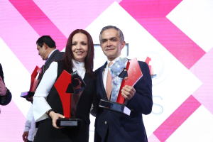 Zwei Personen auf einer Bühne, die lächeln und Trophäen halten, mit einem Bildschirm im Hintergrund