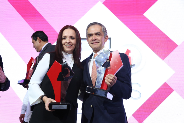 Zwei Personen auf einer Bühne, die lächeln und Trophäen halten, mit einem Bildschirm im Hintergrund