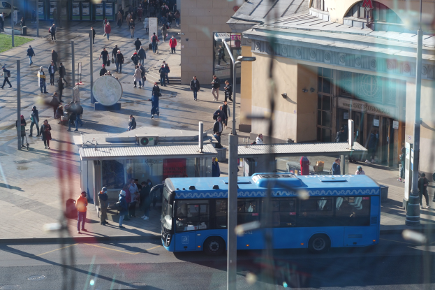 Blauer Bus fährt auf einer Straße mit hohen Gebäuden, Fußgängern, Pfählen und Gras.