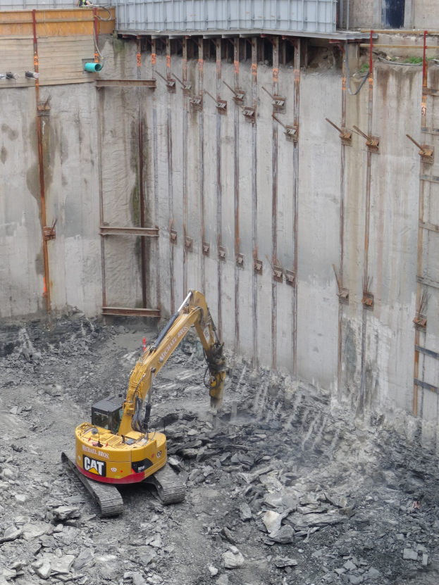 Bagger beim Einsatz auf einer Baustelle mit Steinen und Eisenstangen neben einem teilweise errichteten Gebäude