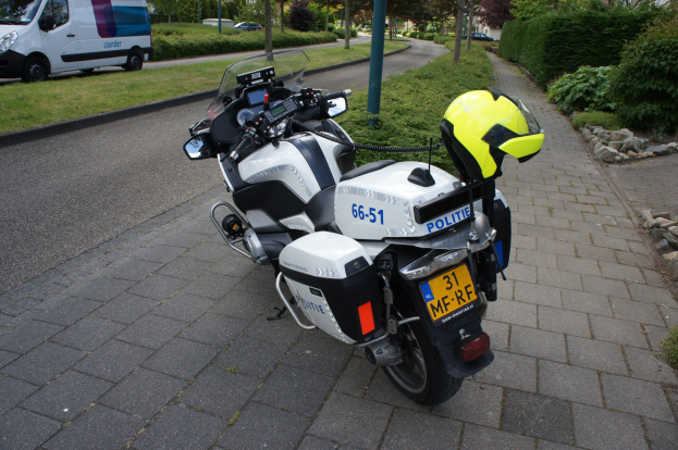 Ein auf der Straße geparktes Polizeimotorrad mit einem Helm darauf, umgeben von Bäumen, Polen, Gebäuden, Pflanzen, Gras, Steinen und anderen Fahrzeugen im Hintergrund.
