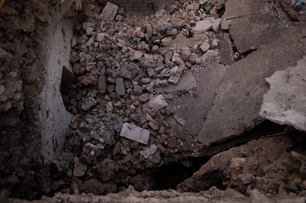 Zusammengebrochenes Gebäude mit einem großen Loch im Boden, umgeben von Trömmern und einer sichtbaren Leitung, die Erdbebenschäden anzeigt.