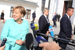 Bundeskanzlerin Angela Merkel spricht vor der Reichstagskuppel mit Reportern, umgeben von einer Gruppe von Menschen, einige halten Mikrofone, mit Gebäuden im Hintergrund.