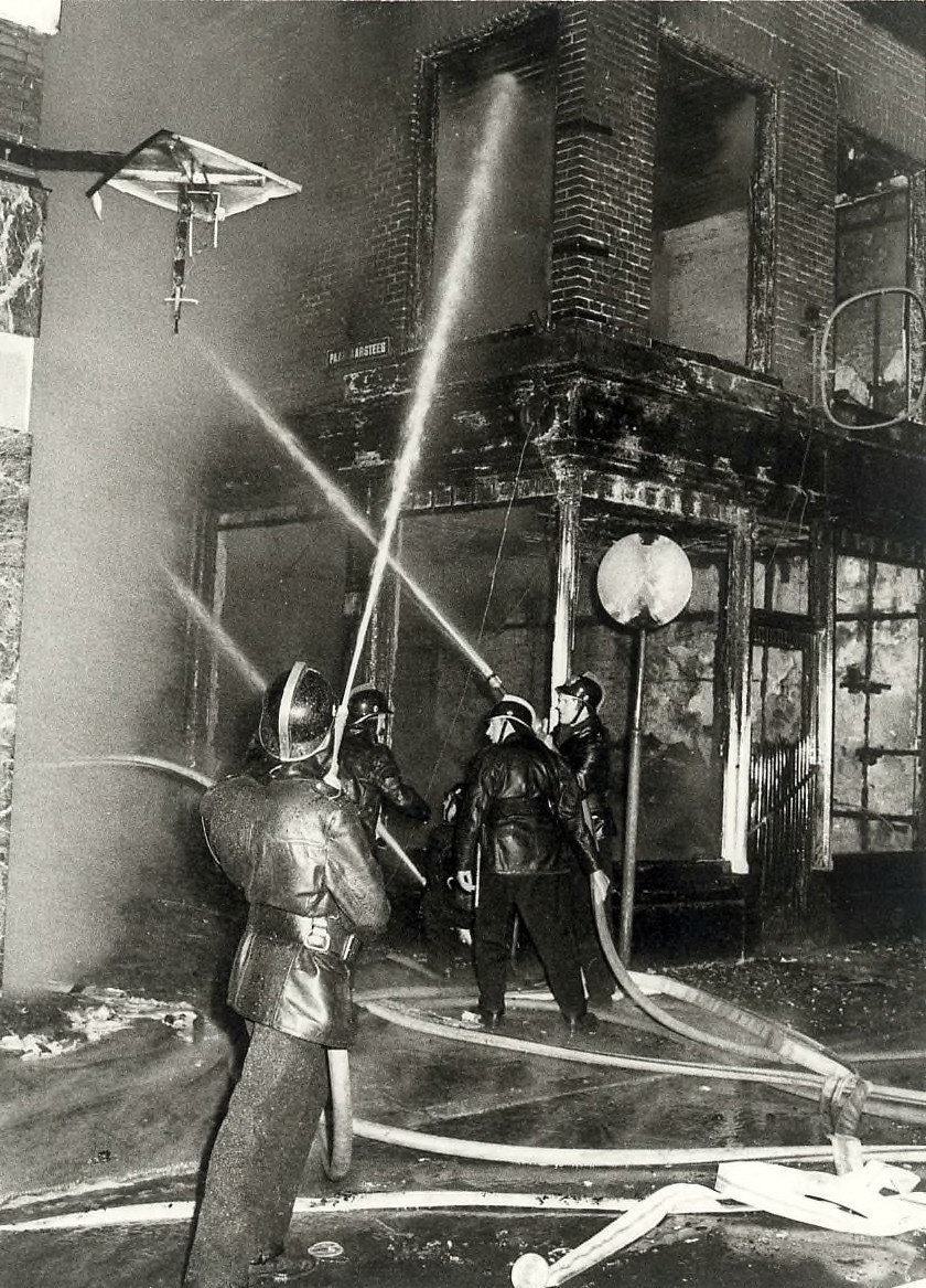 Schwarzes und weißes Bild von Feuerwehrmännern in Helmen mit Schläuchen in den Händen, die vor einem feuerbeschädigten Gebäude stehen.