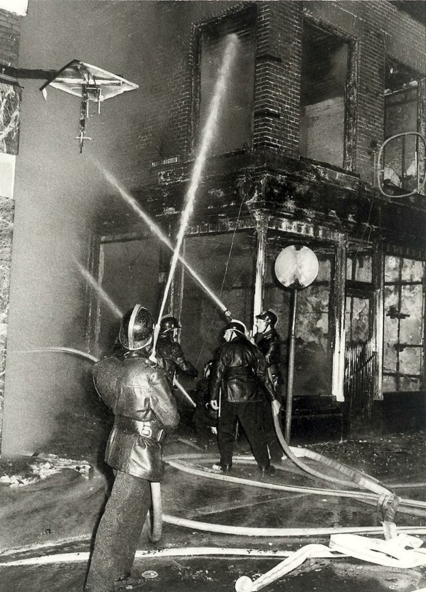 Schwarzes und weißes Bild von Feuerwehrmännern in Helmen mit Schläuchen in den Händen, die vor einem feuerbeschädigten Gebäude stehen.