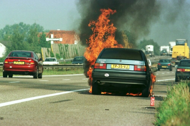 Ein Auto brennt am Straßenrand mit anderen Fahrzeugen in der Nähe, Bäumen und Gebäuden im Hintergrund bei einem klaren blauen Himmel und Gras mit einem Feuerlöscher rechts.