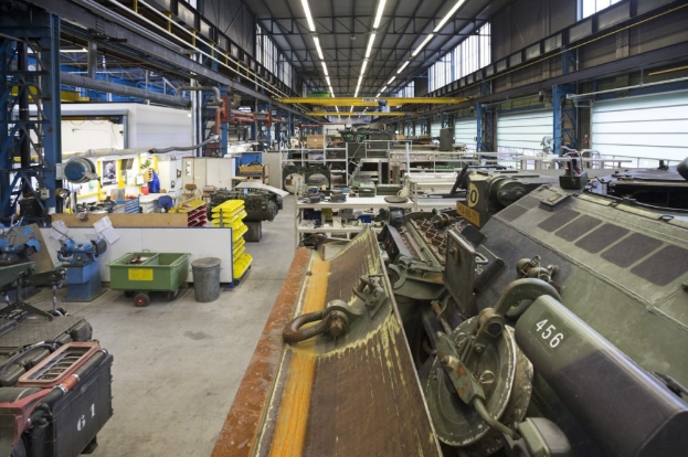 Großes Lager mit Maschinen, ein Landfahrzeug auf der rechten Seite, Metallobjekte auf der linken Seite und Deckenbeleuchtung.