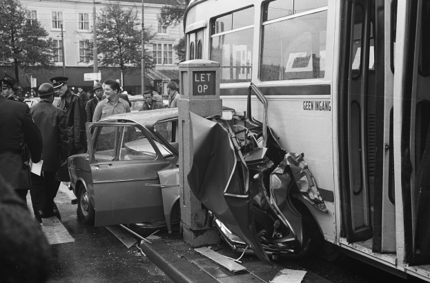 Schwarz-weißes Bild eines Busses, der auf der Seite der Straße in ein Auto gekracht ist, mit Menschen drumherum und Bäumen und Gebäuden im Hintergrund.