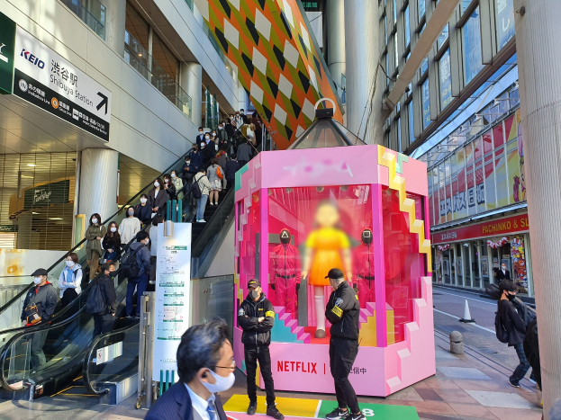 Eine Gruppe von Menschen steht vor einem Gebäude mit Glaswänden, Säulen und Texttafeln, einige tragen Masken, während eine Rolltreppe mit Menschen im Hintergrund fährt; ein pinkfarbener Karton in der Mitte zeigt Schaufensterpuppen und trägt die Aufschrift "Netflix's new Netflix booth in Tokyo."
