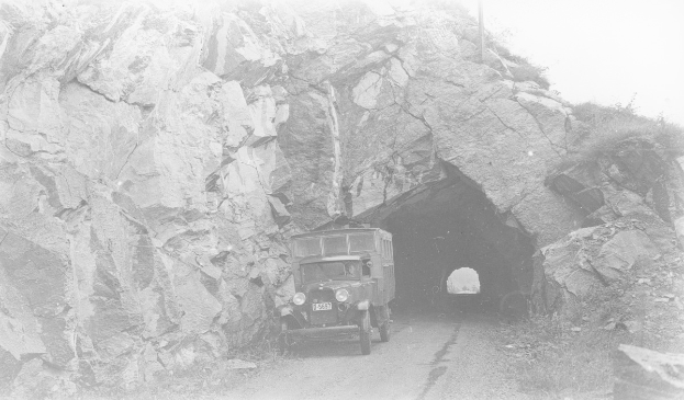 Schwarzes und weißes Foto eines Lkw, der durch einen Tunnel mit felsigen Wänden auf beiden Seiten fährt.
