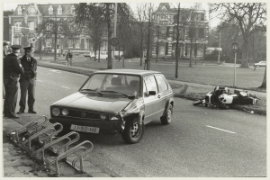 Ein Schwarz-Weiß-Foto eines Autos, das auf der Seite der Straße in ein Motorrad gekracht ist, mit drei Personen auf dem Gehweg links neben dem Auto und Bäumen, Pfählen, Gebäuden und anderen Fahrzeugen im Hintergrund.