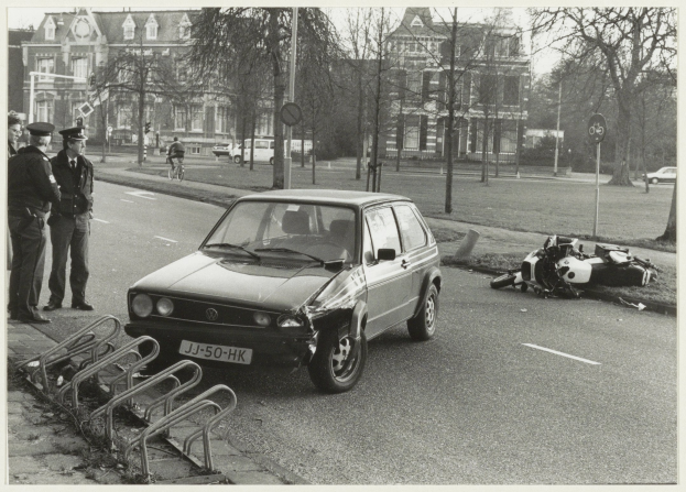 Ein Schwarz-Weiß-Foto eines Autos, das auf der Seite der Straße in ein Motorrad gekracht ist, mit drei Personen auf dem Gehweg links neben dem Auto und Bäumen, Pfählen, Gebäuden und anderen Fahrzeugen im Hintergrund.
