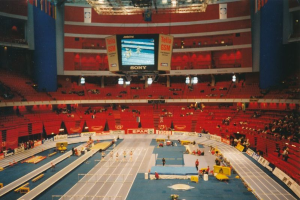 Eine große Halle mit Zuschauern bei einer Leichtathletikveranstaltung, mit Sitzplätzen, Stehplätzen, Texttafeln, Lichtern und einem Bildschirm oben.
