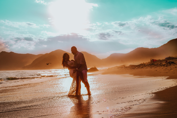 Ein Paar umarmt und küsst sich am Strand bei Sonnenuntergang, mit farbenfrohem Himmel, tiefblauem Wasser, saftig grünen Hügeln und einem Vogel am Himmel.