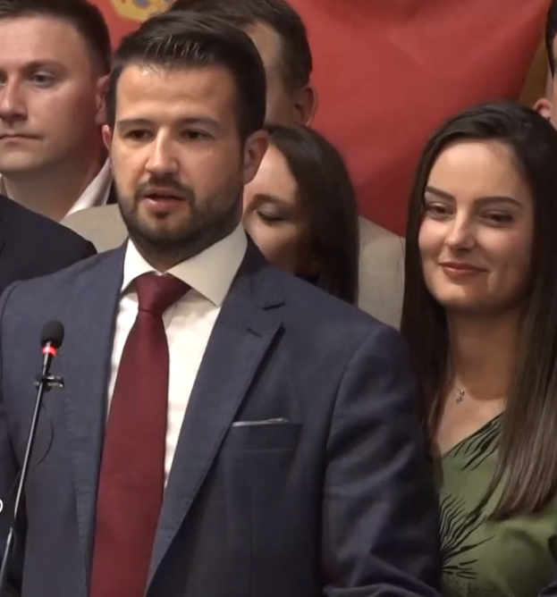 Ein Mann im Anzug und Krawatte steht an einem Podium mit einem Mikrofon und spricht vor einer Menge mit einem Banner im Hintergrund, wahrscheinlich auf eine Regierungssperre für soziale Medien reagierend.