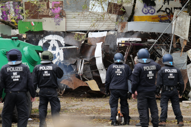 Eine Gruppe von Polizisten in schwarzen Uniformen und Helmen steht vor einem Haufen Müll, mit einem grünen Fahrzeug, einem Gebäude mit Graffiti und einem Baum im Hintergrund.