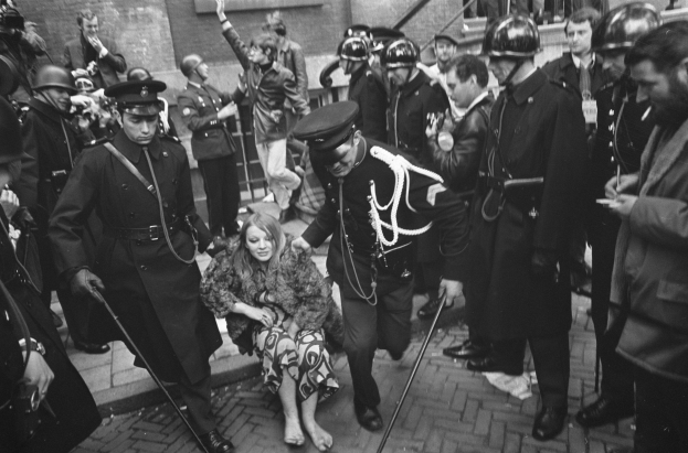 Schwarzes und weißes Foto einer Frau in Polizeigewahrsam, die von einer Menge Helme tragender Menschen mit Stöcken eskortiert wird, mit einem Gebäude mit Treppe und Geländer im Hintergrund.