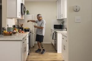 Ein Mann steht in einer Küche mit weißen Schränken, hält ein Glas in der Hand und sieht verschiedene Küchenutensilien auf einer Arbeitsplatte links von ihm.