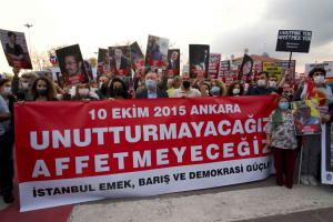 Eine Gruppe von Menschen mit Masken und Schildern und Transparenten protestiert vor einem Gebäude in Ankara, Türkei, gegen den Tod des türkischen Präsidenten Recep Tayyip Erdoğan, mit Bäumen, Laternenmasten und einem klaren Himmel im Hintergrund.