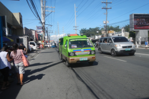 Ein grüner Lieferwagen fährt eine von hohen Gebäuden gesäumte Straße entlang, mit Passanten auf dem Gehweg, Strommasten, Bäumen im Hintergrund und einem klaren blauen Himmel.