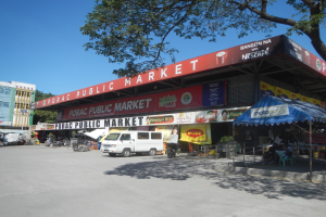 Ein öffentlicher Markt mit einem vor ihm geparkten Van, umgeben von Gebäuden, Geschäften, Namensschildern, Kraftfahrzeugen, Menschen unter einem Zelt sitzend, Geländern, Bäumen und einem klaren blauen Himmel.