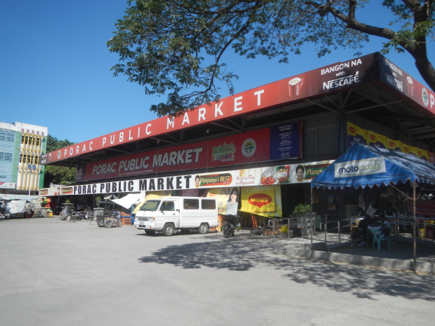 Ein öffentlicher Markt mit einem vor ihm geparkten Van, umgeben von Gebäuden, Geschäften, Namensschildern, Kraftfahrzeugen, Menschen unter einem Zelt sitzend, Geländern, Bäumen und einem klaren blauen Himmel.