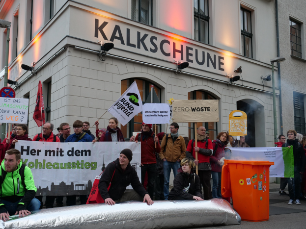 Eine Gruppe von Menschen mit Protestschildern und Plakaten vor einem Gebäude, mit zwei Personen im Vordergrund und einem Müllcontainer rechts, vor einem Hintergrund von Gebäuden mit Fenstern und Schildern in Deutschland.